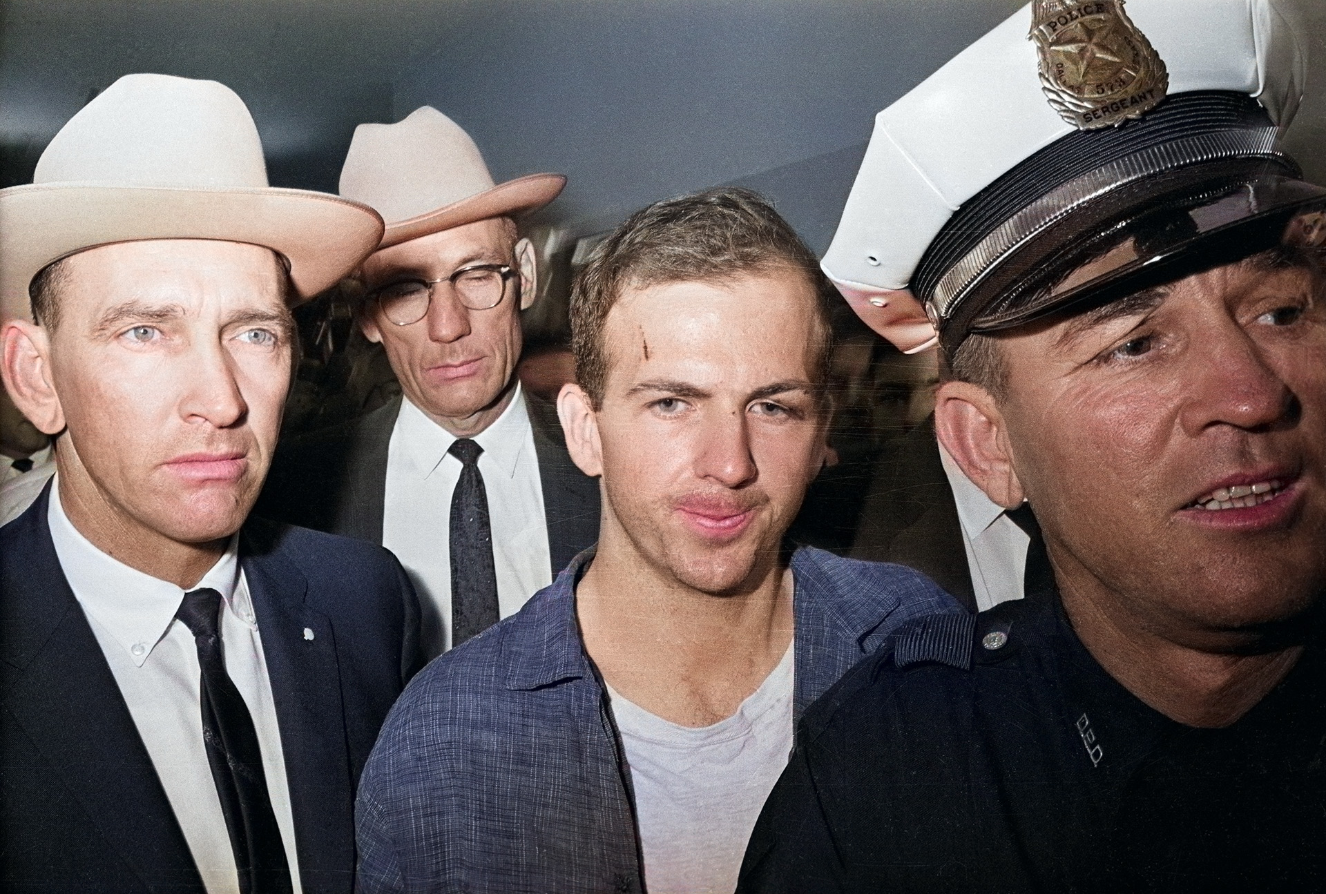 Colorized image of Lee Harvey Oswald in handcuffs, escorted by law enforcement officers.