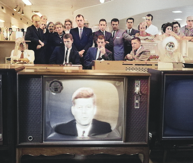 Customers and staff in a 1960s electronics store stop to watch President John F. Kennedy on television as he addresses the nation about the Cuban Missile Crisis.