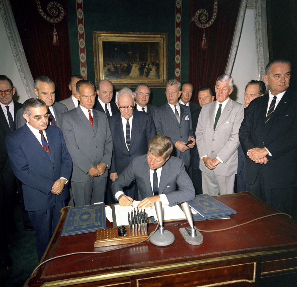 President Kennedy, seated at a desk, signs the ratified Test Ban Treaty, surrounded by a group of lawmakers.