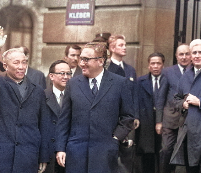 Colorized photo of Dr. Henry Kissinger and Le Duc Tho walking side by side, with Tho waving to onlookers.
