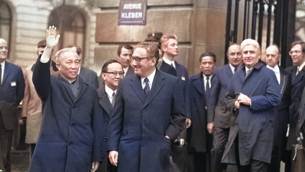 Colorized photo of Dr. Henry Kissinger and Le Duc Tho walking side by side, with Tho waving to onlookers.