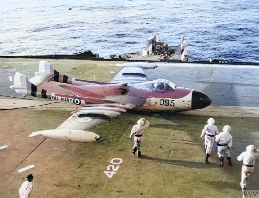 A flak-damaged Sea Venom aircraft with campaign markings on the deck of HMS Eagle, with crew members nearby, following a sortie over Egypt in November 1956.